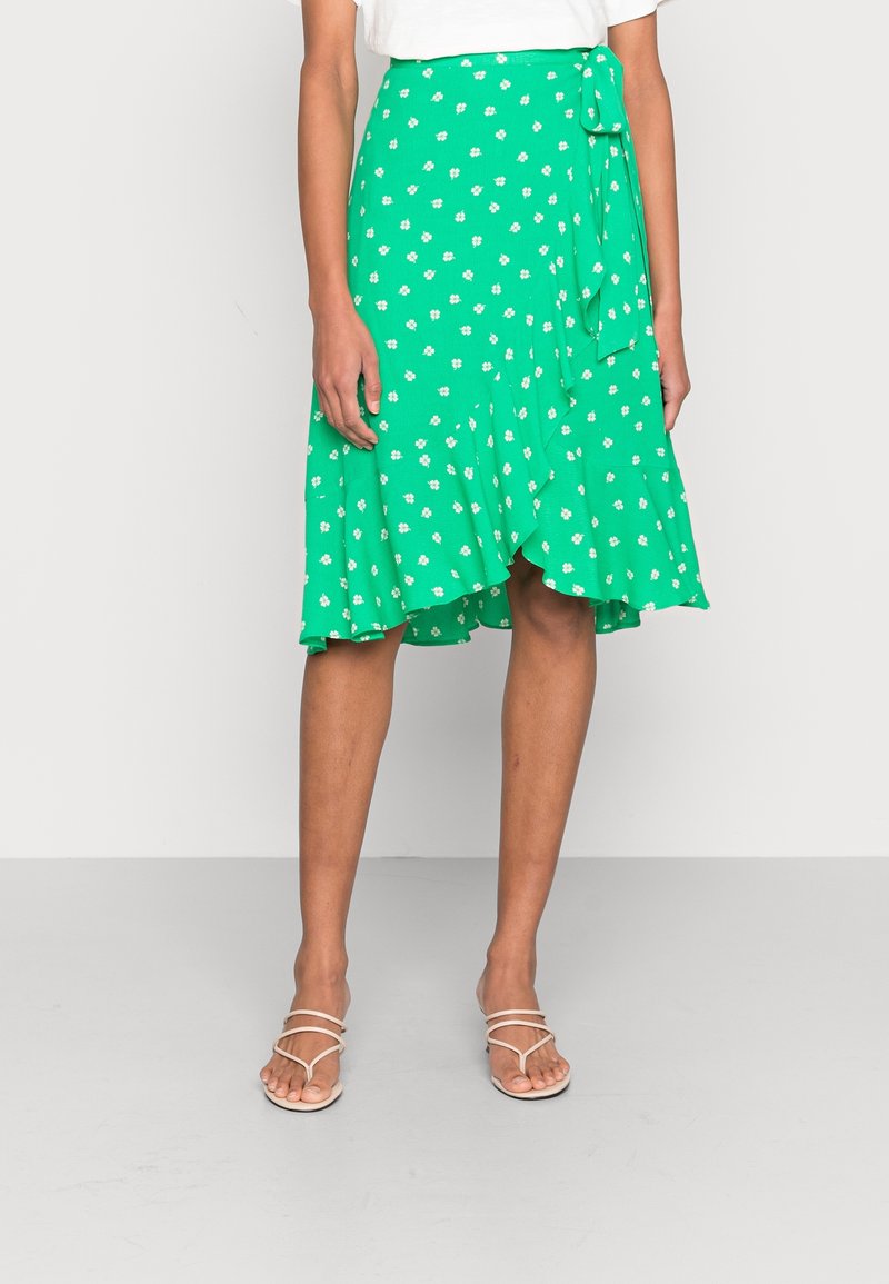 Jupe portefeuille verte à imprimé floral blanc avec lien sur le côté, portée avec des sandales blanches à brides et un haut blanc, montrée de la taille aux pieds.
