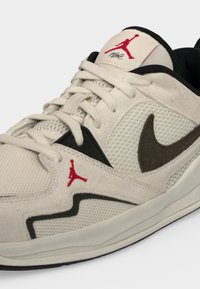 Μπεζ αθλητικό παπούτσι Nike Air Jordan με μαύρο λογότυπο swoosh, κόκκινα λογότυπα Jumpman στη γλώσσα και πλευρά, και μπεζ κορδόνια.