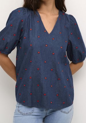 Denimblå bluse med korte pufærmer og rødt hjertebroderi, båret sammen med lyseblå jeans.