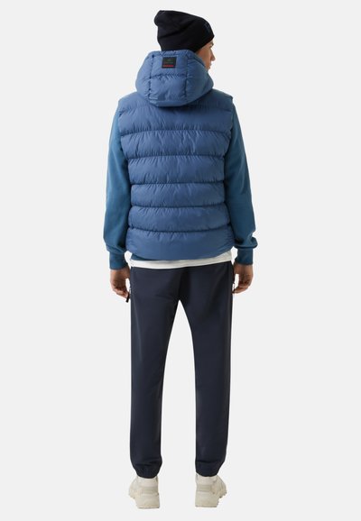 Blaue gepolsterte Weste mit Kapuze, über einem blauen Langarmshirt getragen, kombiniert mit dunklen Hosen und hellen Sneakern.
