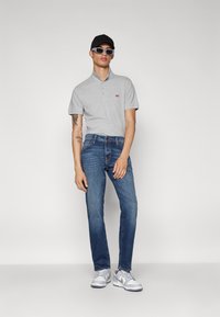 Camisa polo gris con un pequeño logo rojo, jeans de mezclilla azul, zapatillas gris, y una gorra negra; el modelo lleva gafas de sol y está de pie.