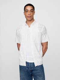 Hvit kortermet skjorte med broderte blomsterdesign, knappestolpe foran og en enkelt brystlomme, båret over en enkel hvit undertrøye.