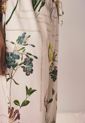 Beežide pükste lähivõte siniste ja lillade lillemustritega ning roheliste lehtedega, rõhutatud beeži nööriga ja osaliselt käega.