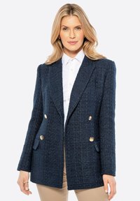 Dunkelblauer, doppelt geknöpfter Blazer aus strukturiertem Stoff, mit goldenen Knöpfen und zwei Vordertaschen, kombiniert mit einem weißen Hemd darunter.