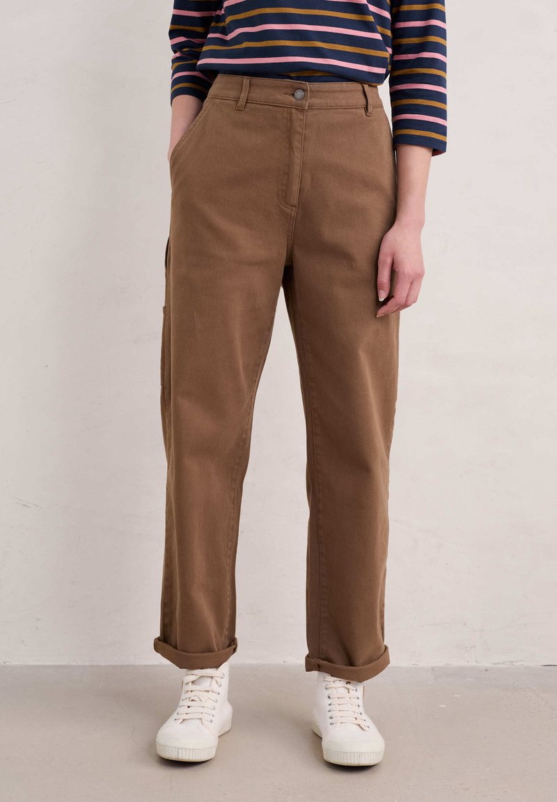 Pantalones de algodón marrón con corte recto, puños enrollados y bolsillos frontales, combinados con zapatillas deportivas blancas y una camiseta de rayas de manga larga.