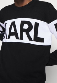 Musta neulepusero, jossa on leveä valkoinen kuvio, jossa lukee suurin kirjaimin "KARL". Pyöreä kaula-aukko ja pitkät hihat, joissa on kontrastiväriset raidat.