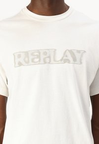 Hvit bomulls t-skjorte med en lys grå "REPLAY" logo i en forvrengt skrifttype, rund hals og korte ermer. Glatt tekstur.