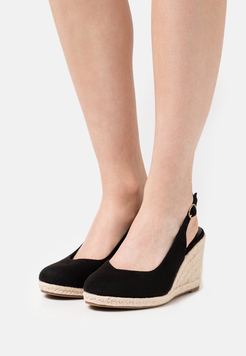 Pieds portant des espadrilles compensées noires à bout fermé avec semelles en corde beige et brides à la cheville sur fond blanc.