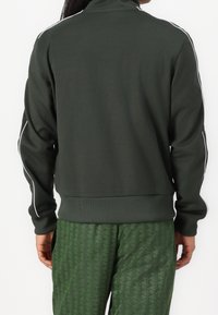 Sudadera con cremallera verde oscuro con rayas blancas a los lados, puños y dobladillo acanalados; combinada con pantalones verdes con estampado de diseño geométrico.
