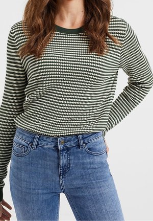 Femme portant un pull à manches longues à motifs verts et blancs avec des poignets et un col vert foncé, associé à un jean en denim bleu taille mi-haute.