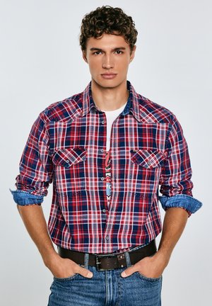 Rød og blåternet skjorte med lange ærmer, to brystlommer og krave i denim, båret over en hvid grafisk t-shirt og blå jeans.