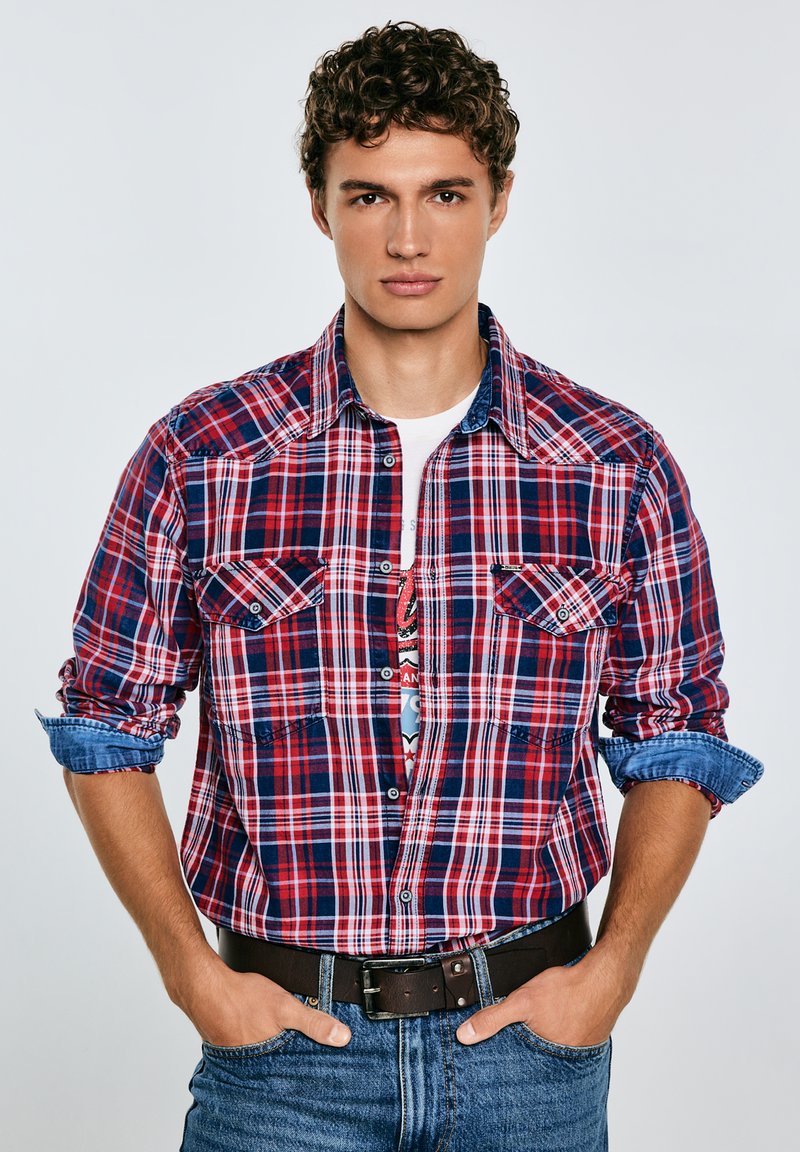 Rød og blåternet skjorte med lange ærmer, to brystlommer og krave i denim, båret over en hvid grafisk t-shirt og blå jeans.