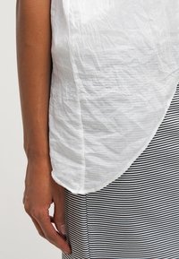 Personne portant une chemise blanche froissée et une jupe rayée noir et blanc, avec la main gauche détendue le long du corps.