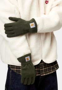 Persona con guantes de punto verde oliva y una chaqueta de forro polar crema sobre una camisa a cuadros y vaqueros oscuros.