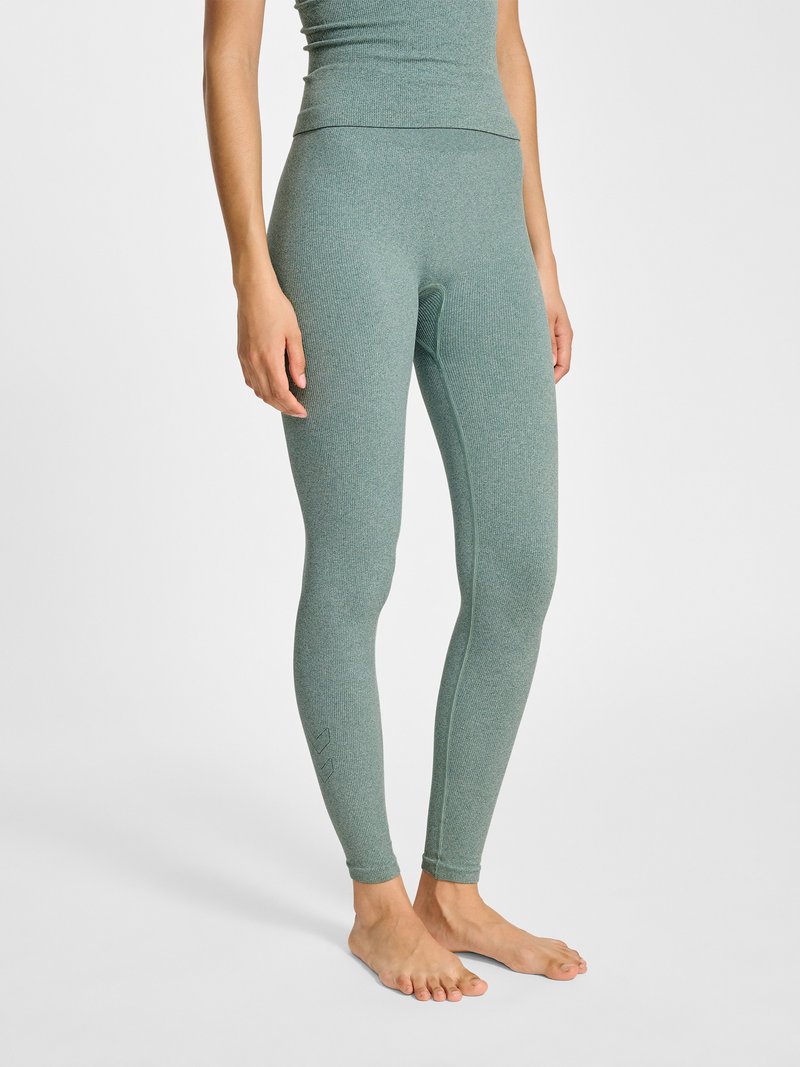 Person trägt eng anliegende, hoch taillierte, strukturierte grüne Leggings und passendes Oberteil, steht barfuß vor einem schlichten weißen Hintergrund.