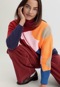 Pull en tricot avec un motif abstrait multicolore dans des tons de rouge, orange, rose, bleu et gris ; manches côtelées ; associé à un pantalon en velours côtelé bordeaux.