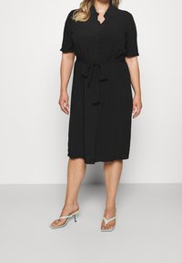 Nainen, jolla on päällä musta nappikiinnitteinen midimekko, jossa on vyötärösolki, ja vaaleansiniset korkokengät, seisoo valkoista taustaa vasten.