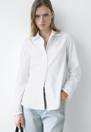 Vrouw met blond haar, een wit doorgestikt hemd en lichte blauwe spijkerbroek, staand tegen een witte achtergrond, met een zwarte riem in haar handen.