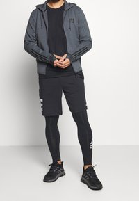 Graue Sportjacke mit Reißverschluss und Kapuze, schwarzes T-Shirt, schwarze Shorts mit weißen Streifen und schwarze Kompressionsleggings mit Logo. Schwarze Sneaker.
