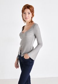 Maglietta lunga a maniche con costine grigia, scollo a V e maniche a campana, con fenditure frontali, abbinata a jeans blu scuro.