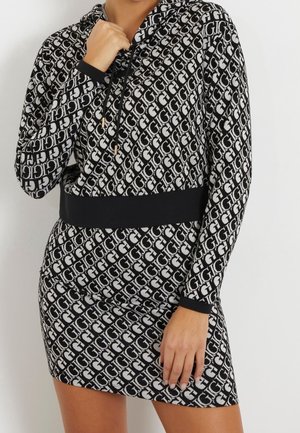 Kvinde iført en sort- og hvidmønstret hoodie-kjole med snører og sort taljebånd, med hånden hvilende ved kraven og låret.