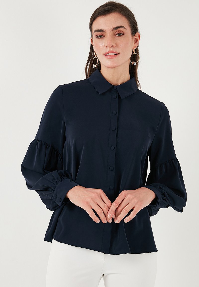 Blusa blu navy con chiusura a bottoni, colletto e maniche a sbuffo. Tessuto morbido e liscio con una vestibilità comoda e dettagli di maniche arricciate.