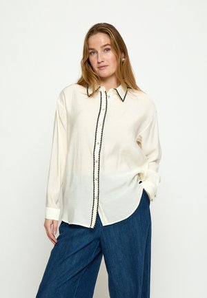 Ženska z dolgimi blond lasmi nosi ohlapno belo srajco z gumbi in črnim robom ter modre široke hlače, pozira z eno roko v žepu.