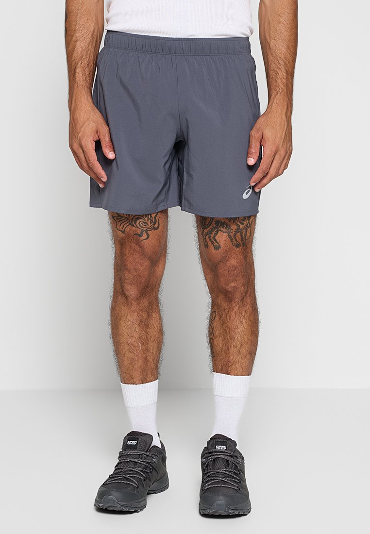 Man in een grijs sportshort, witte kousen tot de enkel, zwarte hardloopschoenen en een witte tanktop, staand tegen een eenvoudige lichte achtergrond.