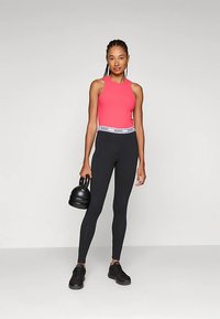 Įliemenuotas rožinis sportinis tankinis marškinėlis ir juodos aukštos liemens tamprės su GUESS logotipo juosta, derinamos su juodais sportbačiais ir laikant kettlebell svarmenį.