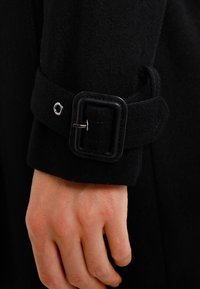 Zwarte wollen jassenmouw met een stoffen band en een rechthoekige plastic gesp, voorzien van een zilveren ring. Hand zichtbaar die de mouw vastpakt.