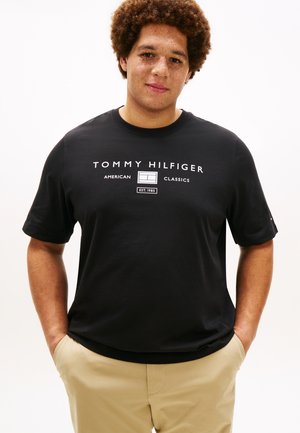 Junger Mann mit lockigem Haar und Sommersprossen trägt ein schwarzes Tommy Hilfiger T-Shirt und beige Hosen mit den Händen in den Taschen vor weißem Hintergrund.