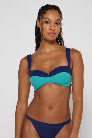 Kobieta z zaplecionymi włosami, ubrana w dwutonowe niebieskie bikini, stoi na jednolitym jasnoszarym tle, patrząc przed siebie z neutralnym wyrazem twarzy.