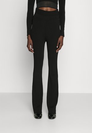 Frau trägt hoch taillierte, gerippte schwarze Schlaghose, kombiniert mit einem kurzen schwarzen Langarmtop und schwarzen Schuhen, steht vor einfachem Hintergrund.