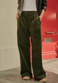 Pantaloni largi din catifea de culoare verde măslin, cu un model în grilă, plisuri și buzunare laterale, asortați cu pantofi slip-on de culoare bej.