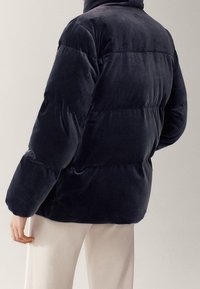 Osoba ubrana w czarną pikowaną kurtkę z poziomym przeszyciem, stojąca z lekko zgiętą lewą ręką, w połączeniu z kremowymi spodniami.