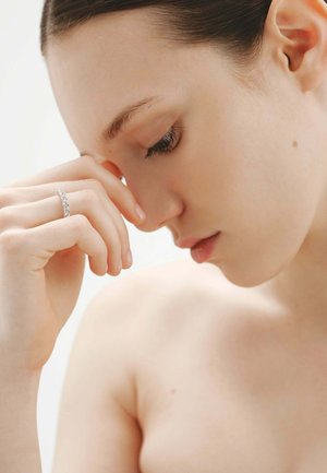 Joven con los ojos cerrados apoyando la frente en su mano, lleva un anillo de diamantes en el dedo, fondo neutro.