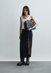 Falda larga negra con cremallera lateral, camiseta blanca sin mangas y bolso de cuero negro. La modelo lleva botas negras gruesas. Diseño minimalista y colores neutros.