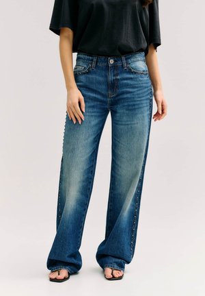 Frau trägt blaue Jeans mit weitem Bein und seitlicher Nietenverzierung, ein schwarzes übergroßes T-Shirt und schwarze Sandalen mit offener Spitze und quadratischem Absatz.