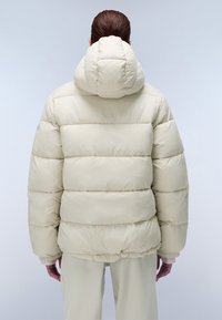 Lichte beige pufferjack met kap, gekenmerkt door een gestept ontwerp, elastische zoom en zachte textuur. Achteraanzicht toont geen extra details.