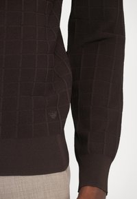 Fekete kötött pulóver, strukturált négyzetmintával, bordás mandzsettákkal és hemekkel, valamint egy diszkrét logó részlettel a deréknál.