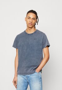 Camiseta de algodón en un color azul marino descolorido, con mangas cortas y cuello redondo. Pequeño detalle de logotipo negro en el pecho. Corte casual.