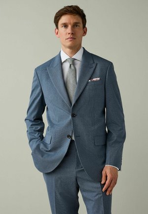 Man in light blue suit, white shirt, patterned grey tie, and floral pocket square stands against plain light background.