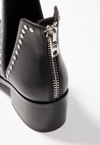Black leather ankle boot with silver studs in a V pattern and a silver zipper on the back heel, standing on a white surface.