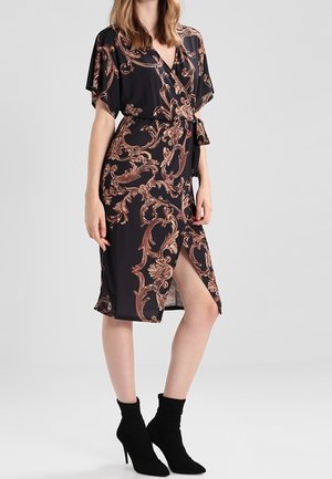 Femme portant une robe portefeuille noire avec des motifs floraux bruns ornés, une ceinture à la taille, et des bottines noires à talons hauts.