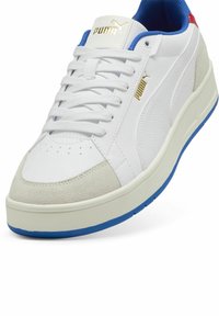 Sneaker bianco con punta in suede, accenti blu sul colletto e sulla suola, logo PUMA in oro, grafica testurizzata sul lato, lacci piatti e suola imbottita.