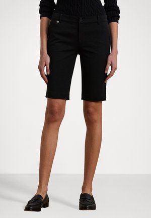 Kvinde iført sorte skræddersyede shorts til knæet, sorte loafers og en sort sweater med kabelstrikkede ærmer.