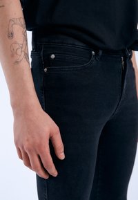 Vaqueros de mezclilla negra con corte ajustado. Incluye dos remaches metálicos, un cierre de cremallera y una textura sutil. El primer plano muestra parte de un brazo con un tatuaje.