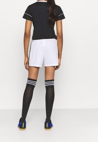 Kvinna stående med ryggen vänd, iförd svart skjorta, vita sportshorts, svarta knästrumpor med vita ränder och blå sportskor.