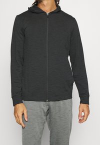Sudadera con cremallera negra y acabado texturizado, con capucha ajustable, combinada con pantalones de chándal grises. Mangas largas y corte relajado.