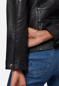 Schwarze Lederjacke mit enganliegendem Ärmel und Druckknopfdetail, kombiniert mit blauen Jeans. Die Textur wirkt glatt und poliert.